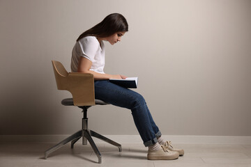 Young woman with bad posture reading book while sitting on chair near grey wall. Space for text - obrazy, fototapety, plakaty