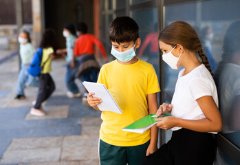 School tweens in medical face masks standing with workbooks in schoolyard during break in lessons. Concept of back to school after lockdown