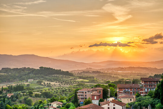 Sunset in small town village of Chiusi Tuscany Italy with houses roof rooftops on mountain countryside rolling hills landscape and orange yellow colorful evening sky with sun rays - Powered by Adobe