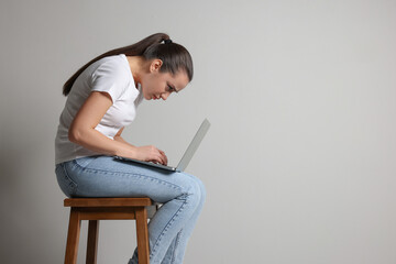 Young woman with poor posture using laptop while sitting on stool against grey background, space for text