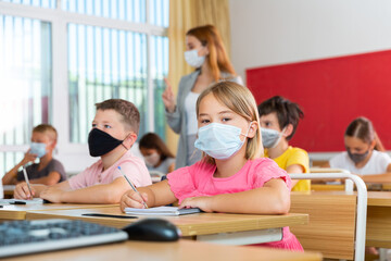 Schoolchildren in masks sitting at desks in classroom. Female teacher staning beside desks.