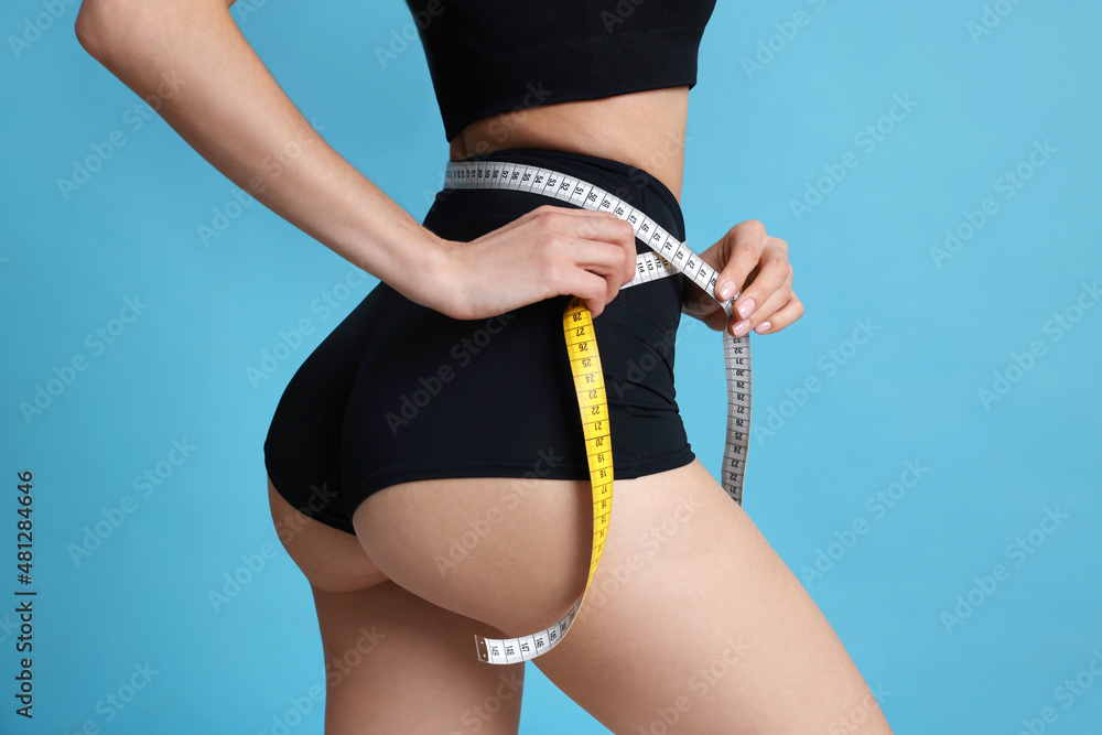 Wall mural Young woman measuring waist with tape on light blue background, closeup