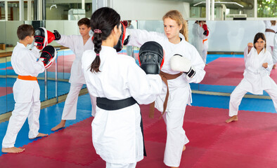 Young girls and boys in boxing gloves exercising jabs during group karate training. Trainer with...