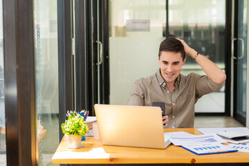 A young businessman is so happy that he uses his smartphone and laptop to analyze financial graphs on his office desk.