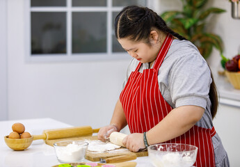Asian young chubby down syndrome autistic autism girl pastry chef model wearing red apron standing threshing pulling bakery flour dough in glass bowl with hands and wooden rolling pin in kitchen