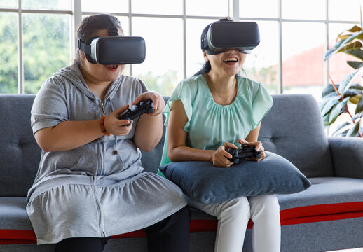 Asian happy lovely mother and young chubby down syndrome autistic autism little daughter sitting on cozy sofa wearing VR virtual reality goggles smiling holding joysticks playing console video game