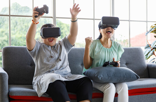 Asian happy lovely mother and young chubby down syndrome autistic autism little daughter sitting on cozy sofa wearing VR virtual reality goggles smiling holding joysticks playing console video game