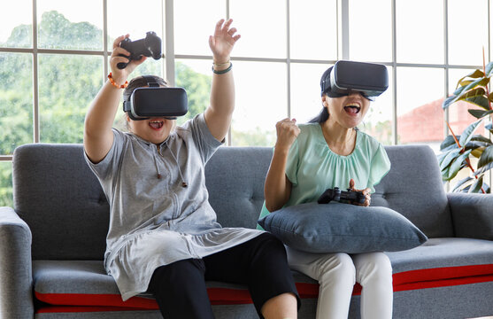 Asian happy lovely mother and young chubby down syndrome autistic autism little daughter sitting on cozy sofa wearing VR virtual reality goggles smiling holding joysticks playing console video game