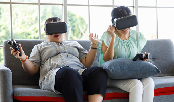 Asian happy lovely mother and young chubby down syndrome autistic autism little daughter sitting on cozy sofa wearing VR virtual reality goggles smiling holding joysticks playing console video game