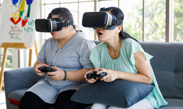 Asian happy lovely mother and young chubby down syndrome autistic autism little daughter sitting on cozy sofa wearing VR virtual reality goggles smiling holding joysticks playing console video game