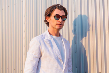 Portrait of a handsome man dressed in a white suit and round hippie sunglasses standing near the...