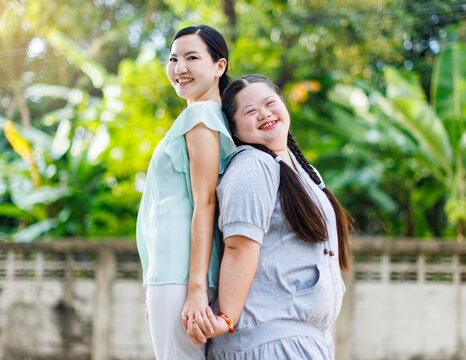 Portrait shot of Asian mother and young chubby down syndrome autistic autism little cute girl with braid pigtail hairstyle stand hugging cuddling embracing together with love smiling look at camera