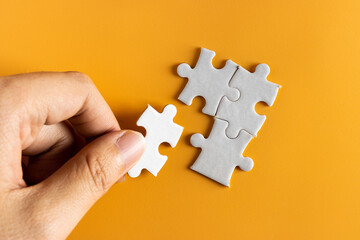 top view of hand holding a piece of jigsaw puzzle. white jigsaw puzzle on yellow or orange...