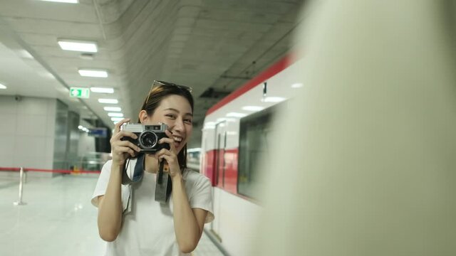 Young beautiful Asian female tourist taking photo with film camera with male friend, smile and enjoyment at train station platform, happy travel lifestyle by subway transport vacation trip.