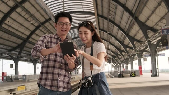 Young couple Asian tourists search information, find travel locations by tablet map at a train station junction in Thailand, passenger holiday trip lifestyle, casual transportation, journey vacation. 