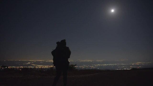 Woman with a tripod and backpack walking under the moonlight, city light in the background. Dolly in. 
