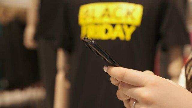 Young woman standing in a shopping mall with a smartphone. Black Friday at the mall. T-shirt black friday on a mannequin. Sales in stores. Search for information in the phone