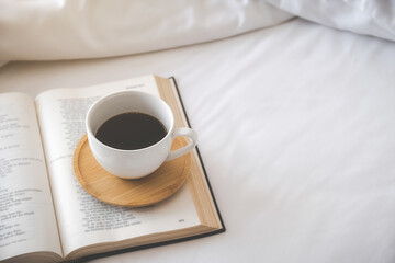 A cup of coffee on book on white bedding sheets in bedroom. Morning lifestyle concept.