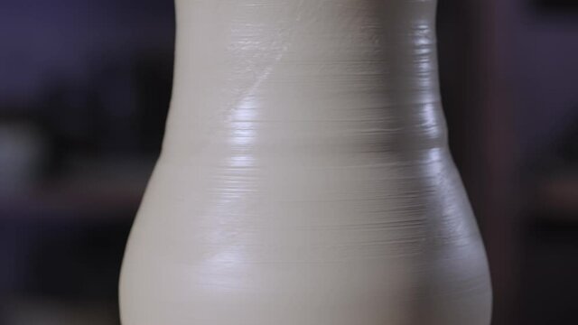 A shaped piece of soft white clay pottery, spinning on a potter's wheel in a creative workshop. A pot molded by the hands of a potter. Earthenware close up in soft light. Slow motion.