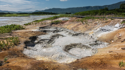 Hot mud springs in the caldera of an extinct volcano. Gray clay, sulfurous orange deposits on the soil. A mountain range against a blue sky. Kamchatka. Uzon