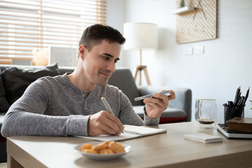 Smiling young man talking on mobile phone and making notes on notebook.