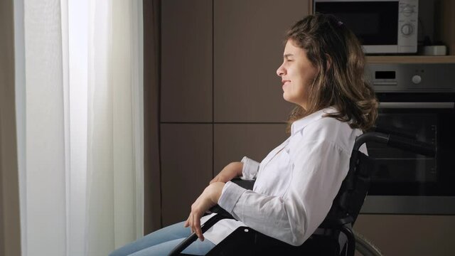 Young disabled woman with organic damage of central nervous system sits in wheelchair looking out of window behind curtain at home side view.