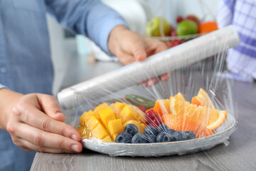 Woman putting plastic food wrap over plate of fresh fruits and berries at wooden table indoors,...