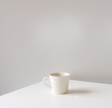 White cup with teabag on edge of table against neutral wall background - minimalist concept