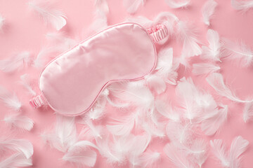 Top view photo of light pink satin sleeping mask and pink feathers on isolated pastel pink background with copyspace