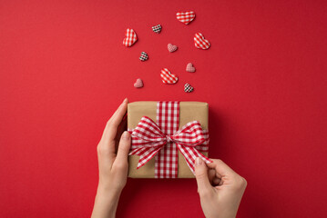 First person top view photo of valentine's day decorations girl's hands unwrapping craft paper giftbox with checkered ribbon bow and hearts on isolated red background