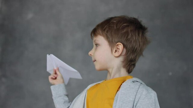 Joyful dreamy boy launches a paper plane into free flight on gray background.