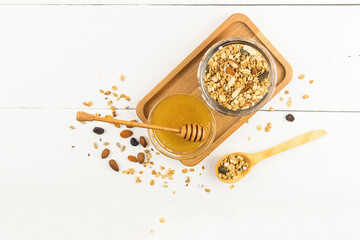 a wooden tray with a glass jar of baked granola with nuts and a bowl of honey. healthy birding. top view. white background.