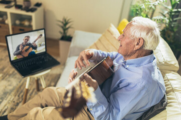 Senior man playing guitar at home using laptop for online lessons - obrazy, fototapety, plakaty