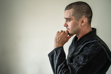 Side view of adult male athlete bjj brazilian jiu jistu black belt standing in front of white wall in kimono gi with copy space looking to the side thinking contemplation