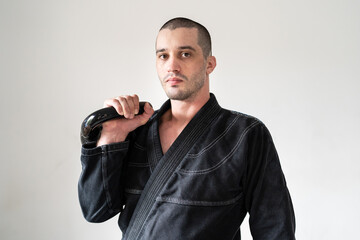 Front view of one man brazilian jiu jitsu bjj athlete standing in front of white wall holding kettlebell weight wearing black kimono gi strength and endurance training concept for martial arts