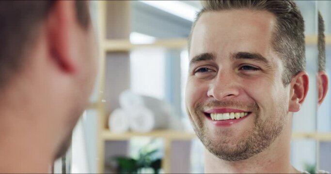 Handsome and happy young man touching his beard in the bathroom mirror at home. Reflection of a confident man smiling as he scans his face and skin, satisfied with a beauty product