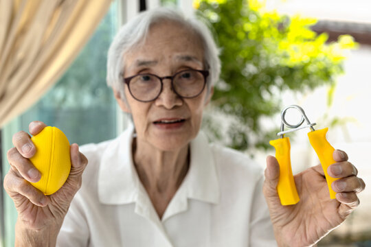 Asian senior woman doing hand,finger and wrist exercise with rubber ball and spring hand grip,stretch finger or squeeze with hand expander,physical therapy or recovery after injury,health care concept - Powered by Adobe