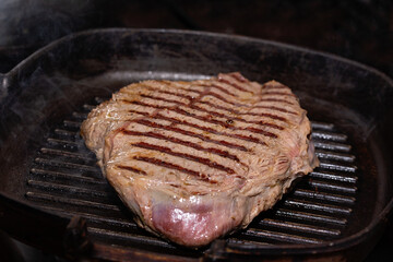 Close-up meat of beef steak grilled in cast-iron ribbed fry pan on gas stove. Delicious juicy meat steak cooking on grill.