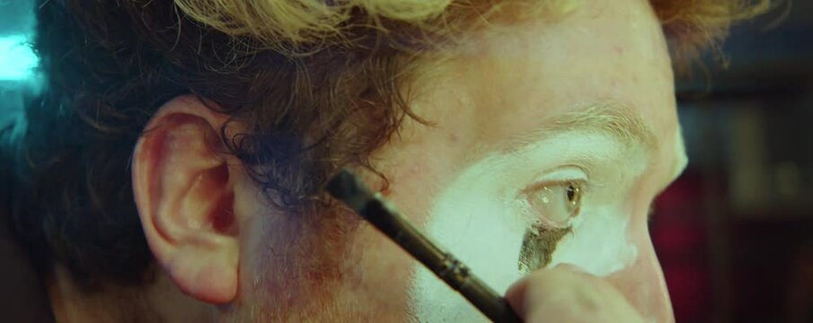 Close up view of male clown applying face paint with brush while doing makeup before performance
