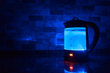 Transparent electric kettle with boiling water. Blue light in dark kitchen.