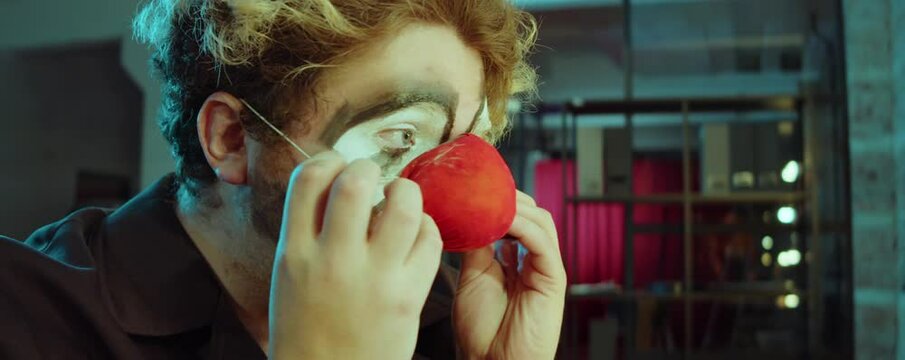 Male Actor With Clown Makeup Putting On Red Nose And Making Faces In Dressing Room