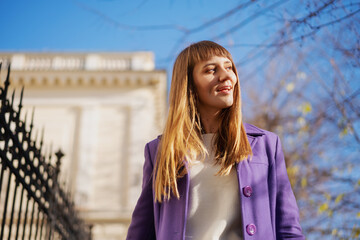 Portrait of a young blonde happy woman in the city in the sunshine in a purple coat