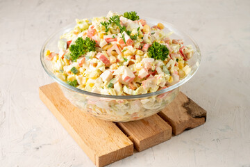 Salad of crab sticks and vegetables in a transparent large bowl on a wooden board, white table.