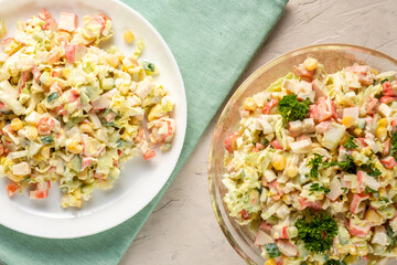 Homemade salad of crab sticks, cabbage, corn, served in a light plate and a transparent bowl, stands on a blue cloth, top view.