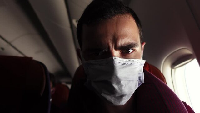 Portrait of angry man in facial mask frowning eyebrows during the flight in the jet. Closeup view of irritated male passenger of airplane sitting next to the window and looking at camera. Bad service.
