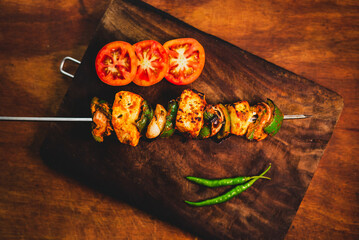 Flat lay of BBQ paneer tikka on a skewer kept on a dark wooden plate with vegetables and fresh tomatoes. Marinated spicy Indian cooked cottage cheese barbequed on fire