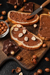 Wooden board of bread with chocolate paste and hazelnuts on table, closeup