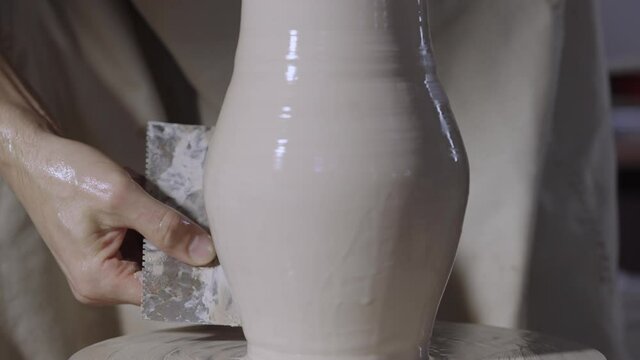 Male hands of potter form a soft white clay pot using pottery tools, turning it on a potter's wheel in a creative workshop. The master gives a shape to the pottery. Clay shaping close up. Slow motion.