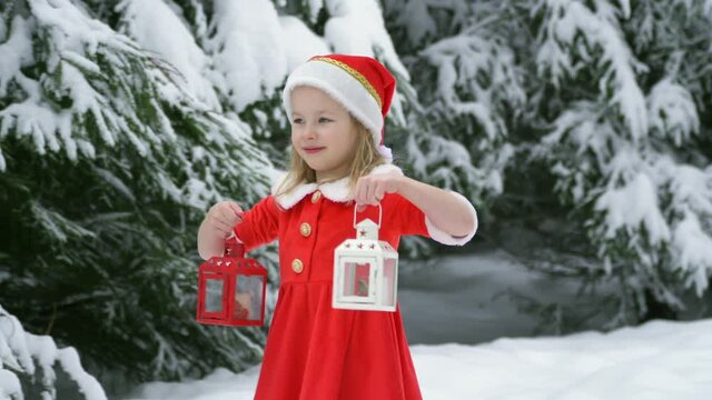 Happy christmas girl smille in santa costume in snow. lamp and candlestick with candles in hands. Winter forest with snowdrifts and snacks. child laughs at new year and walks outside. red suit hat. 