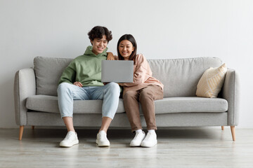 Fototapeta na wymiar Loving millennial Asian couple watching video on laptop, enjoying spare time, sitting on couch at home, copy space